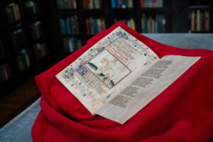 An illustrated manuscript lies open on a piece of red fabric shoing handwritten script and an illustration of a person on a horse.