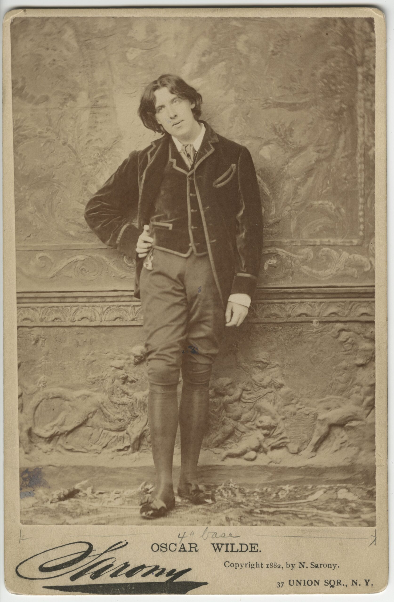 A sepia photo of Oscar Wilde in pants and a jacket with his hand on his hip standing in front of a decorative wall