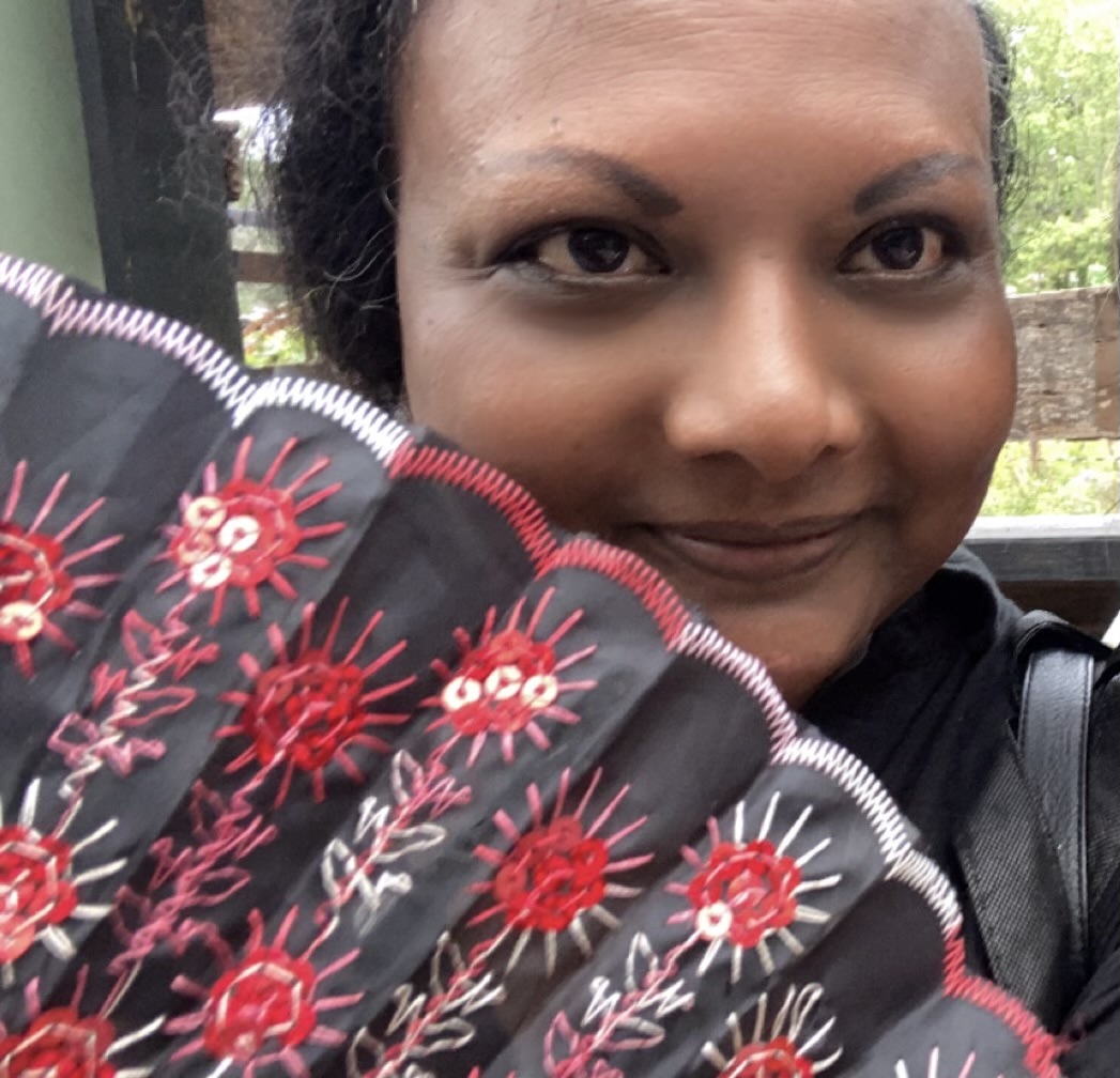 A photo of Sheila Sandapen, a black woman smiles while holding a large red and black fan to her face