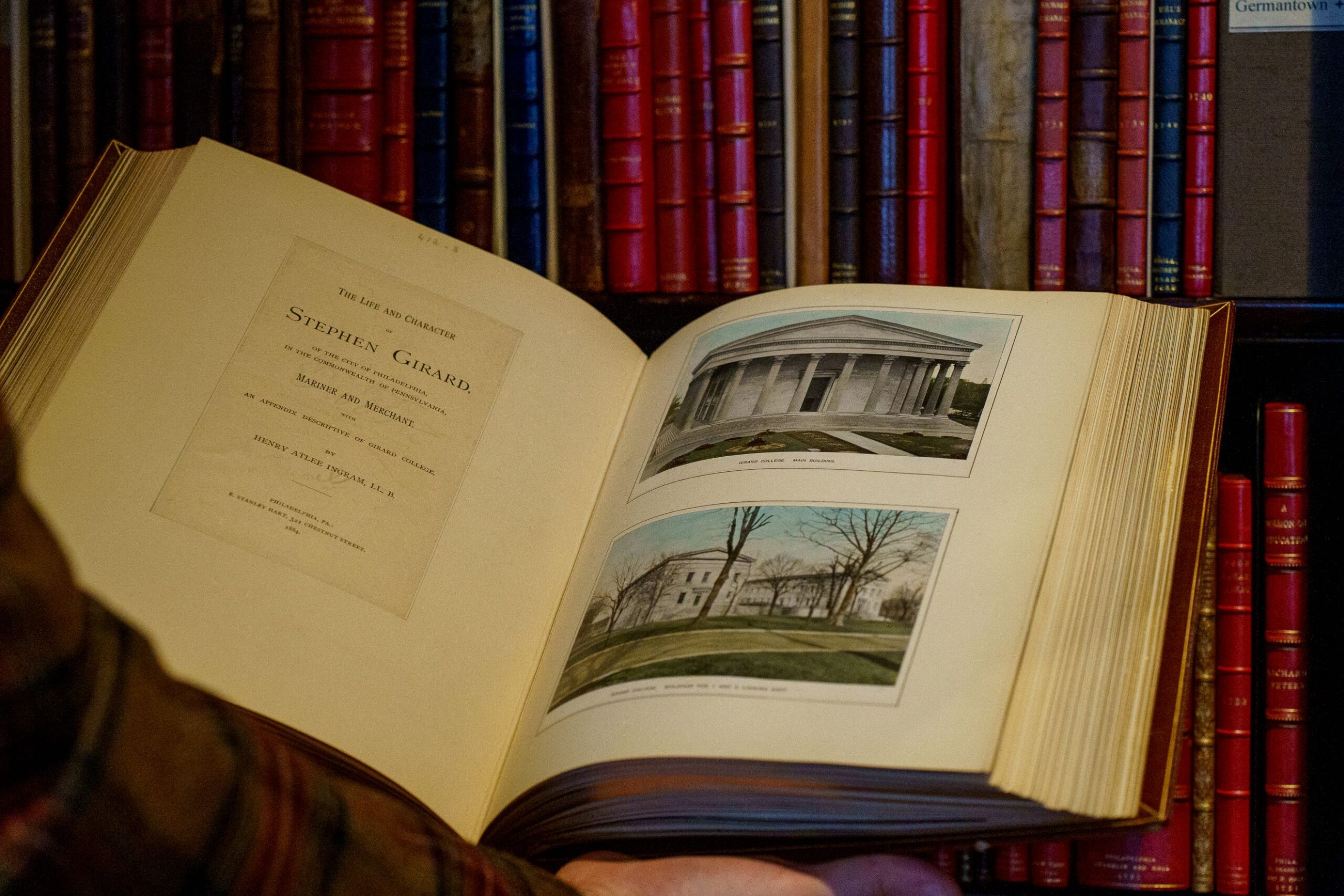 Hands hold an open book revealing illustrations of a large house with white marble columns opposite text that reads Stephen Girard