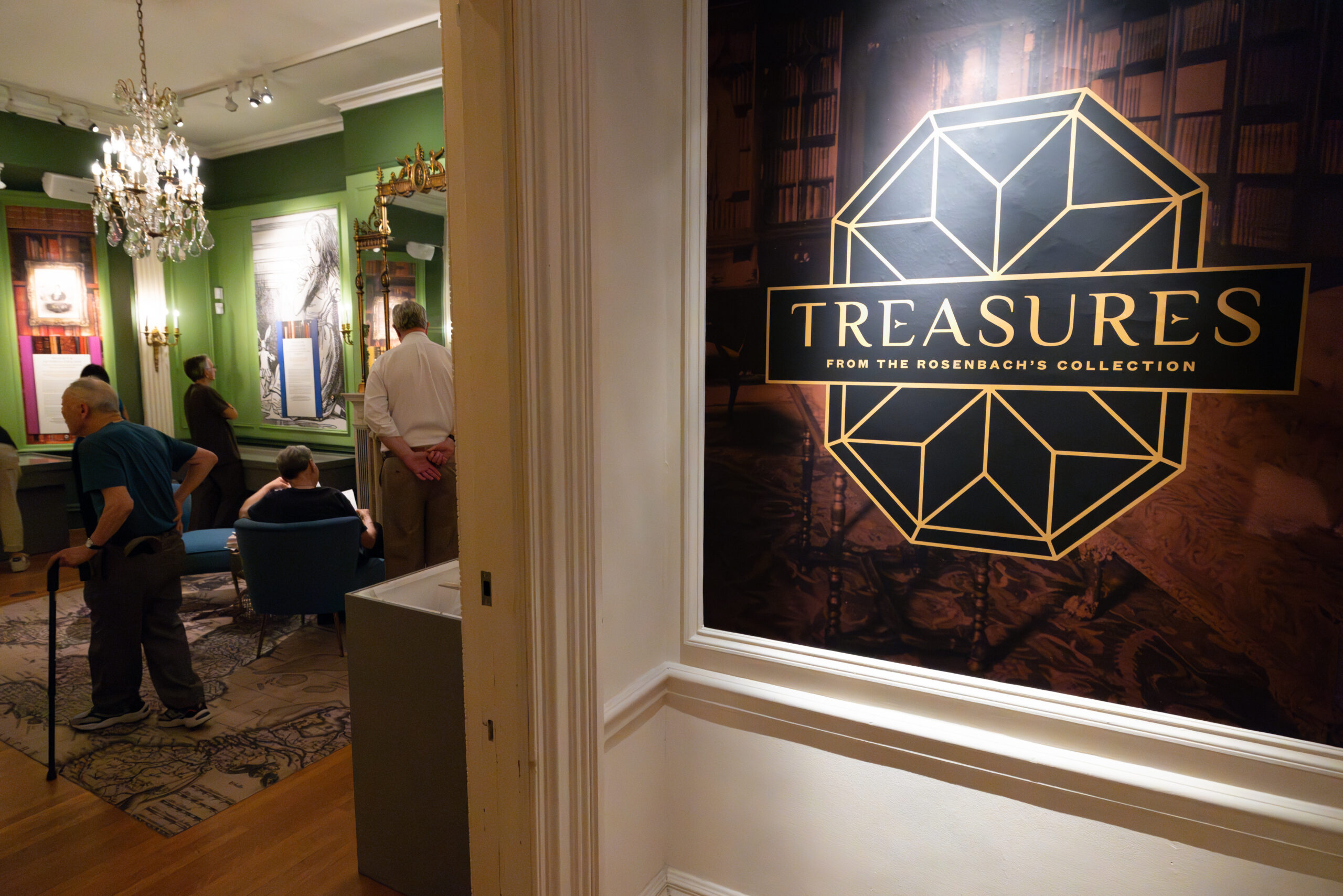 Looking through a doorway into a green gallery space with plexiglass displays, a large brown and gold sign by the door reads "Treasures from the Rosenbach's Collection"