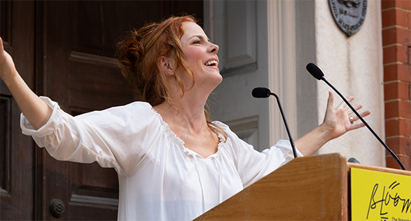 Aa woman in a white night gown smiling and raising her arms as she speaks into a microphone